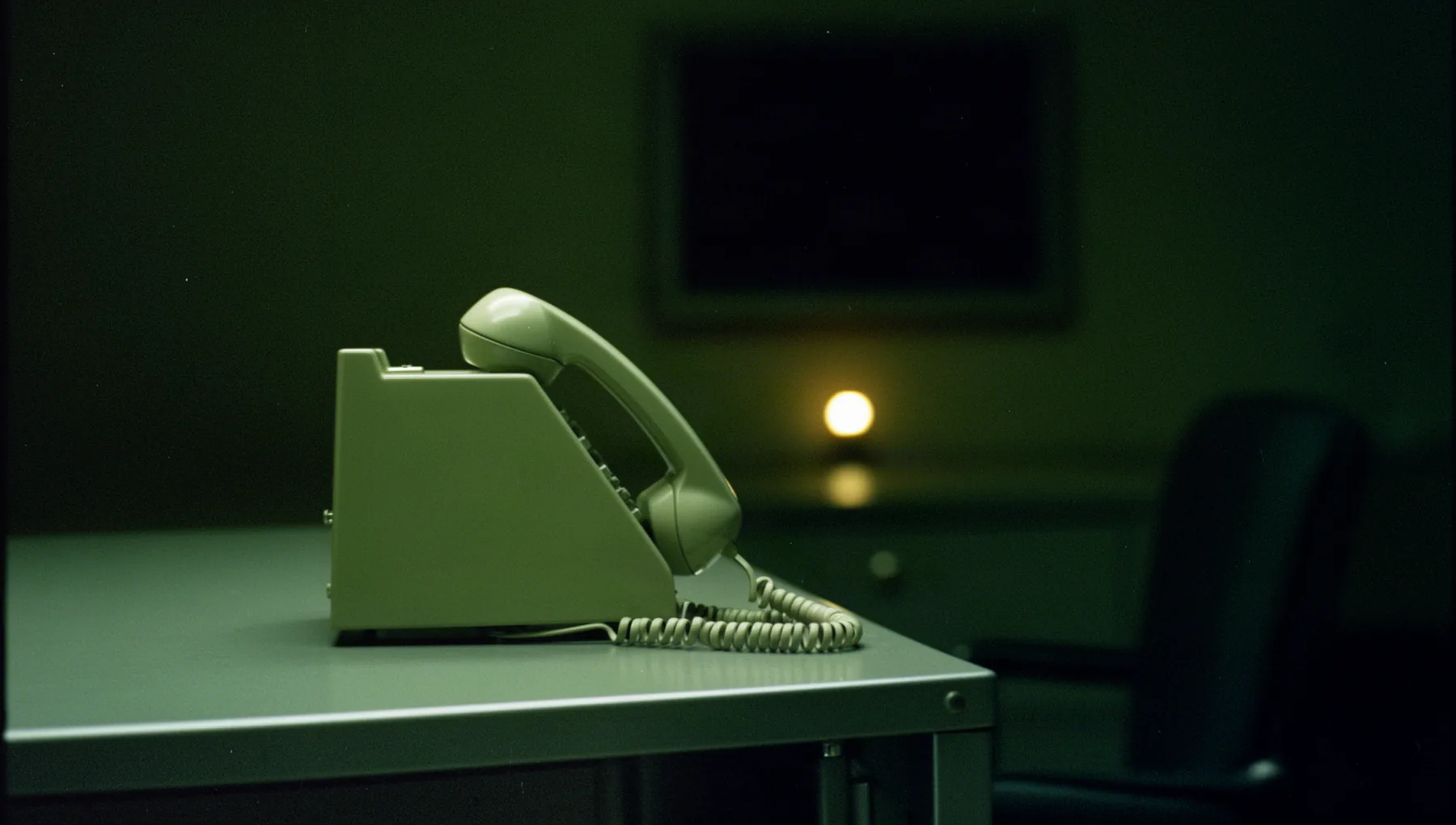 A vintage desk telephone resting in its cradle on an empty steel desk at night, one warm lamp glowing behind it and an empty chair in shadow.