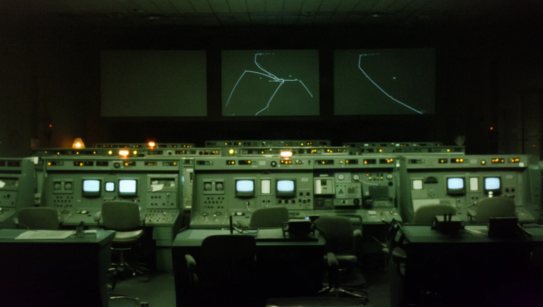 A dark operations floor in a 1969 film look: tiers of steel consoles with small glowing screens and amber lamps, empty chairs, and three large wall screens with faint plotted curves.