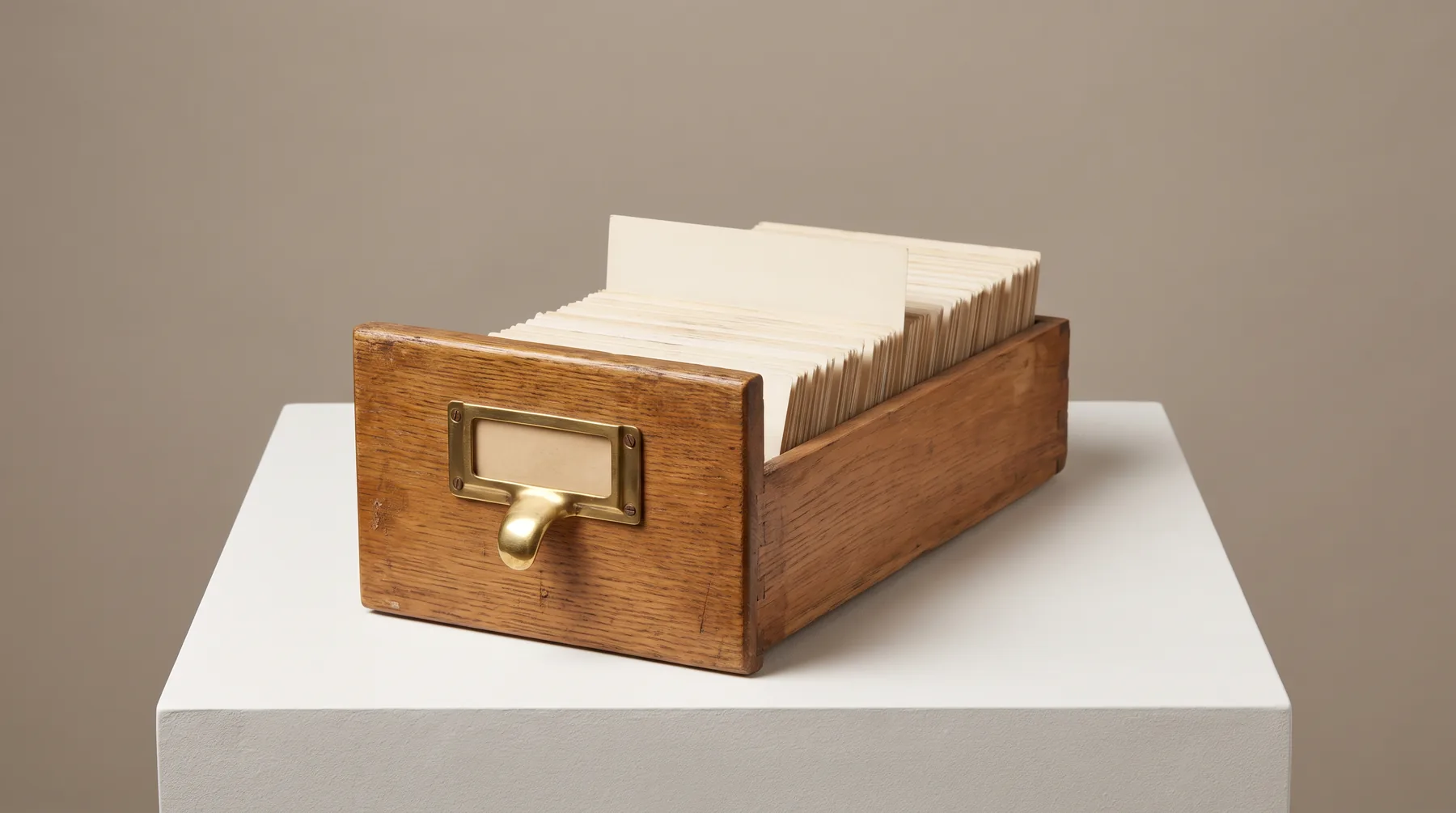 An open oak card-catalogue drawer with a brass handle and an empty brass label frame, full of blank cards with one raised above the rest, on a white plinth.