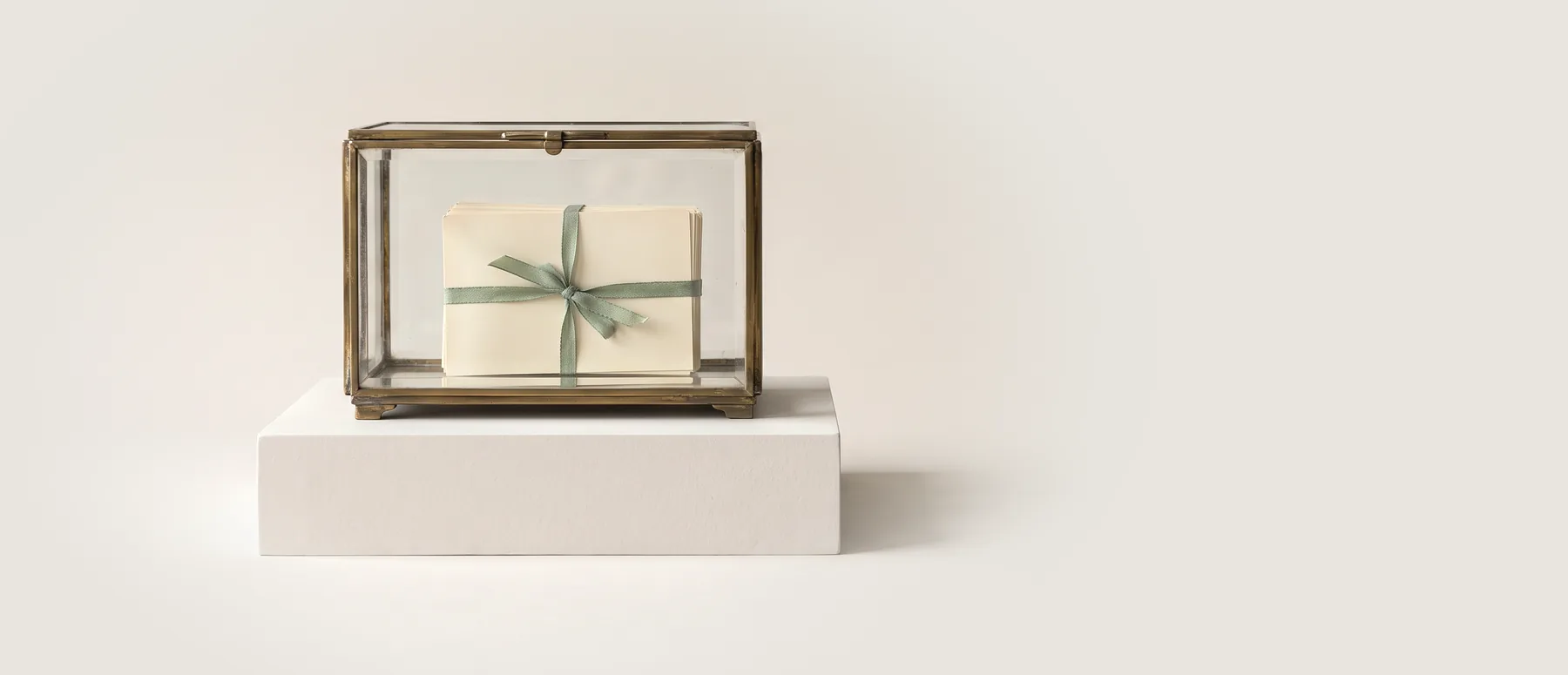A brass-framed glass case on a low white plinth, holding a stack of blank index cards tied with a pale green ribbon, photographed against grey seamless paper.