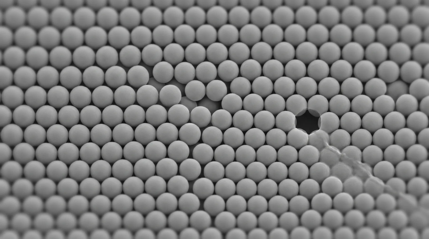 A grayscale, electron-microscope-style field of identical packed spheres with a single sphere missing and a faint dislocation line.